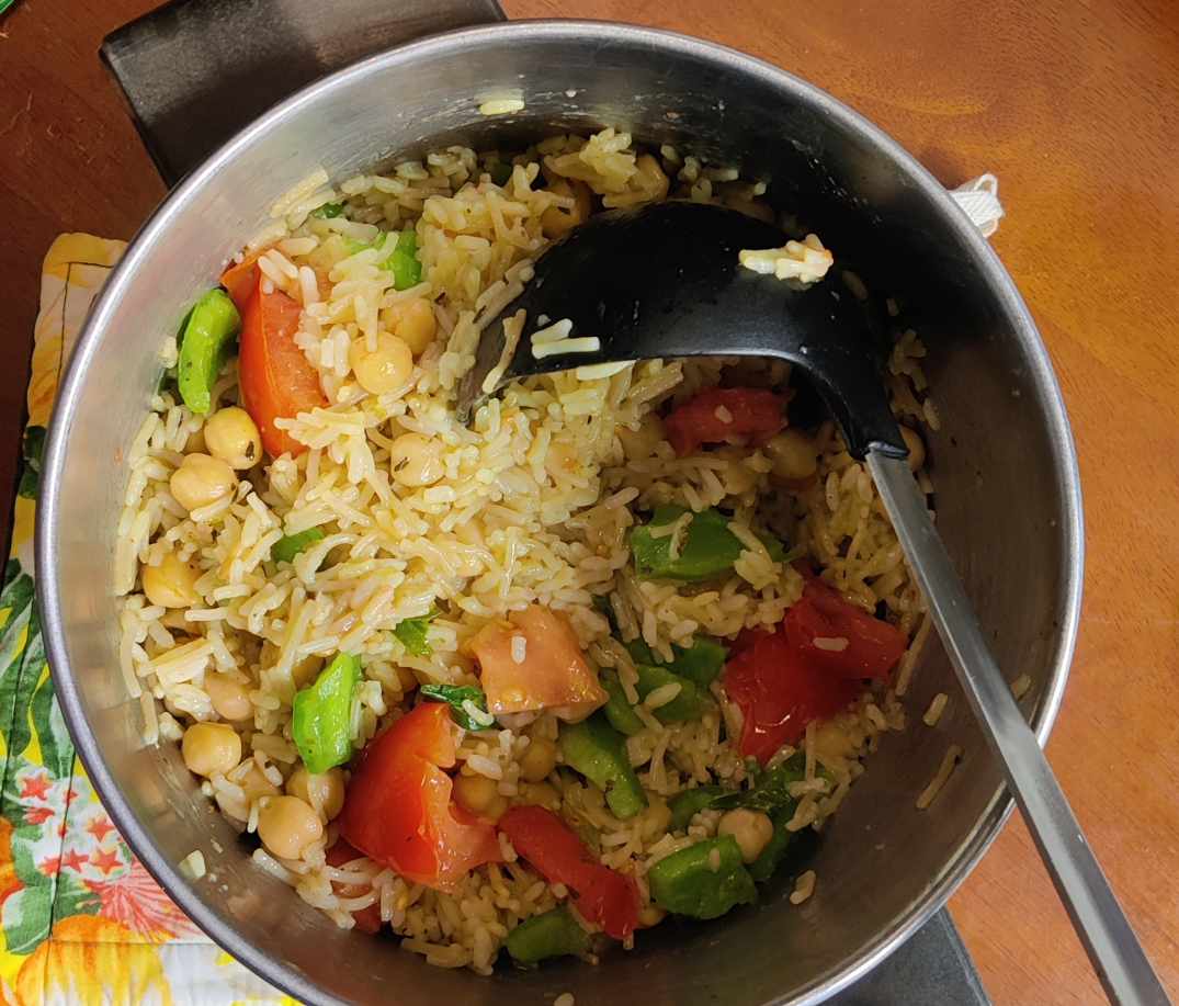 A pot of loaded rice sitting on a floral hot pad. The rice is pale yellow, with chickpeas and chunks of green bell pepper and red tomato mixed throughout.